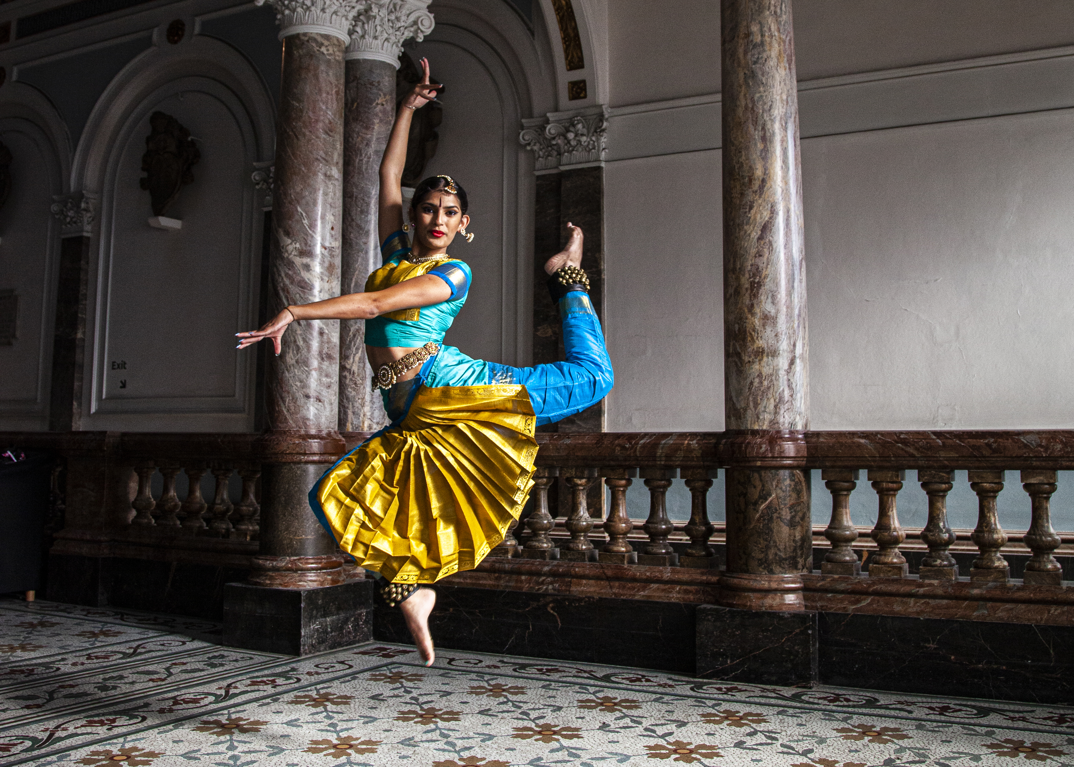 South Asian dancer wearing traditional blue and gold trousers and cropped top with bells on the ankles and a gold jewellery headpiece. Jumping in the air with point toe and one arm above the head and the other crossed over to the left. In front of the museum marble staircase.