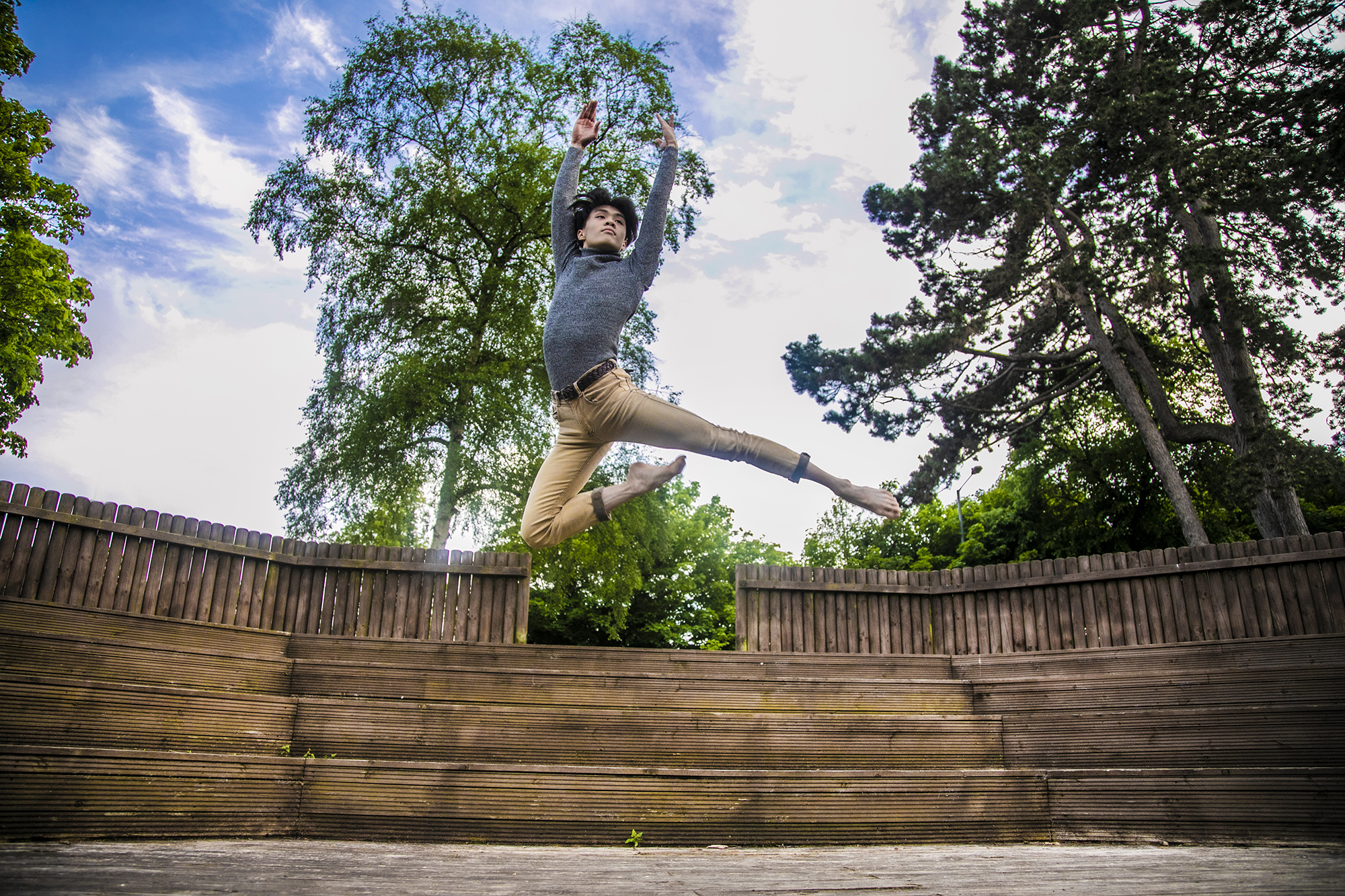 global majority male dancer jumping high with arms above head. Wearing grey long sleve tshirt and beige trousers.  In front of soft focus trees and blue sky.