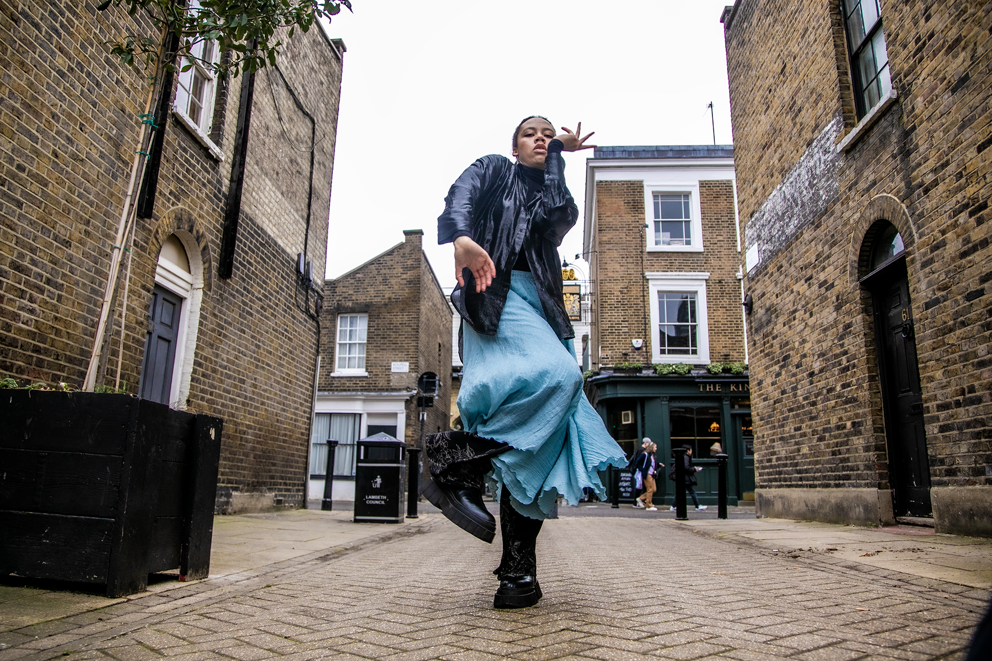 Global majority female dancer dancing down london street facing camera one arm near the face and on one leg . Wearing black leather jacket and blue skirt