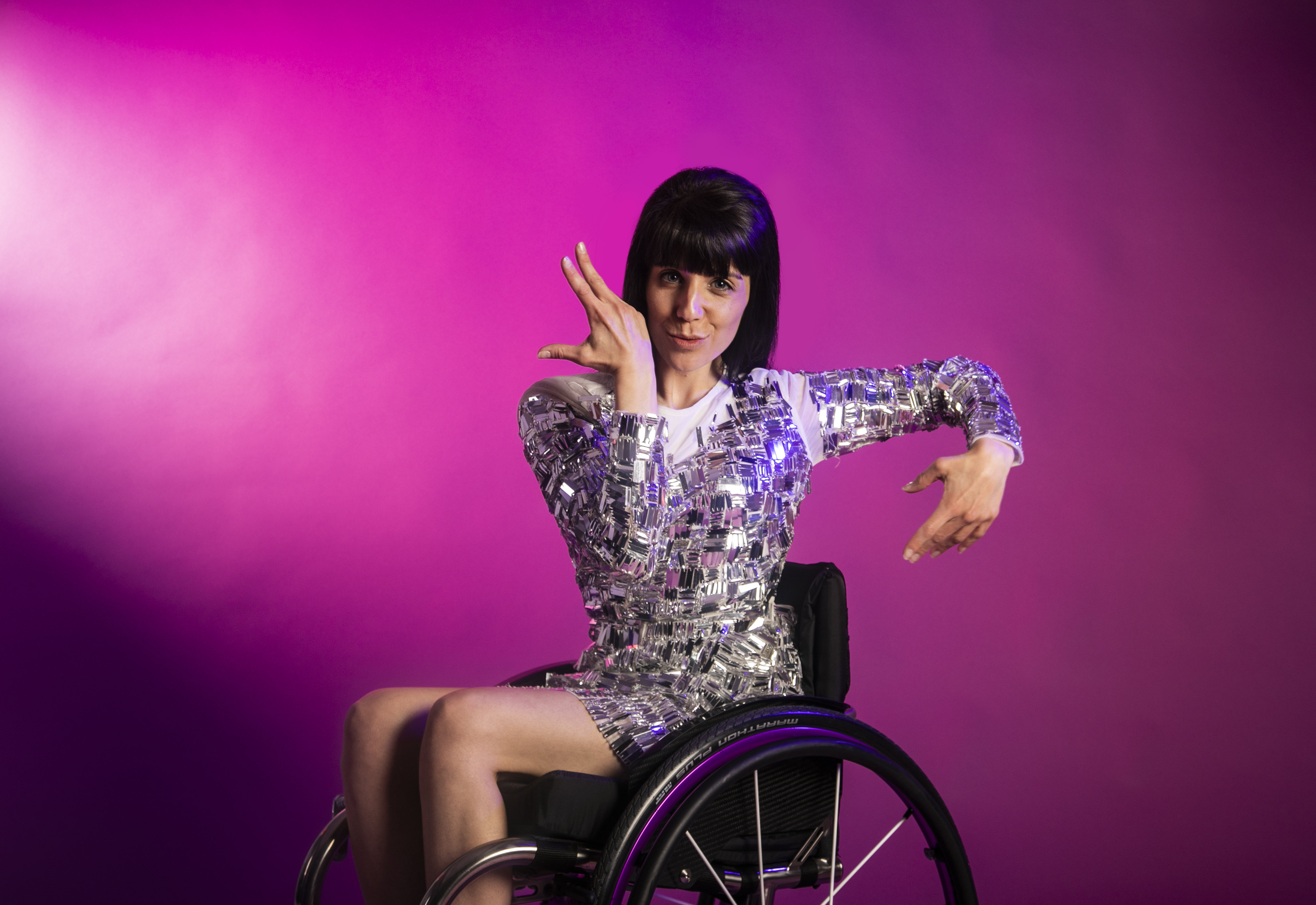 white brunette female dancer in a wheelchair wearing silver sparkly dress. Both hands in salsa movement around her face. On a pink photo backdrop