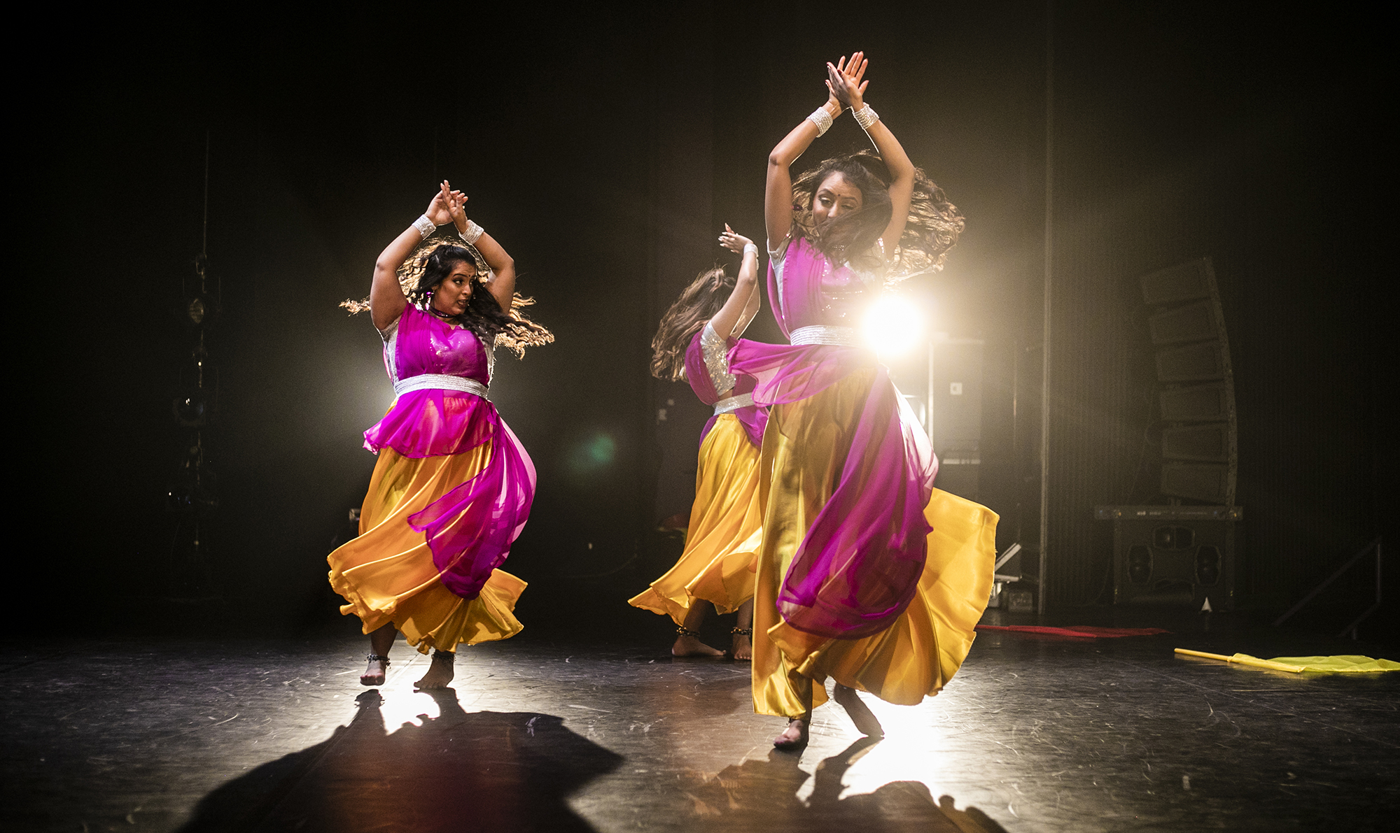 Three female Bollywood dancers spinning on a dark stage wearing pink and yellow sari dresses.