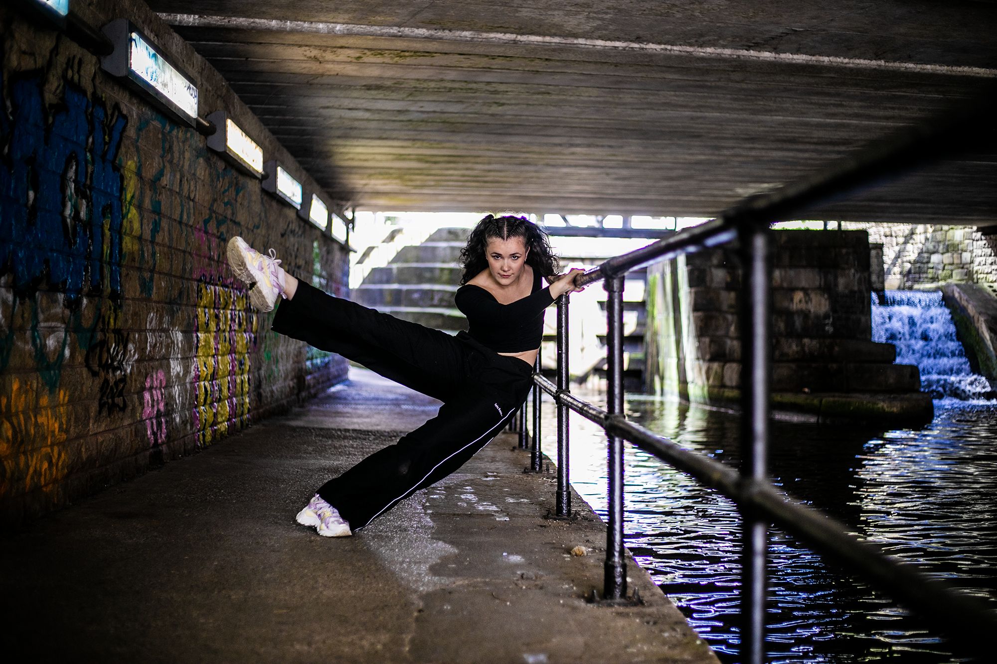 white female dancer with brown curly hair leaning on a raining undernieth a tunnel with water to the right side of the image. wearing blacktracksuit and white trainers