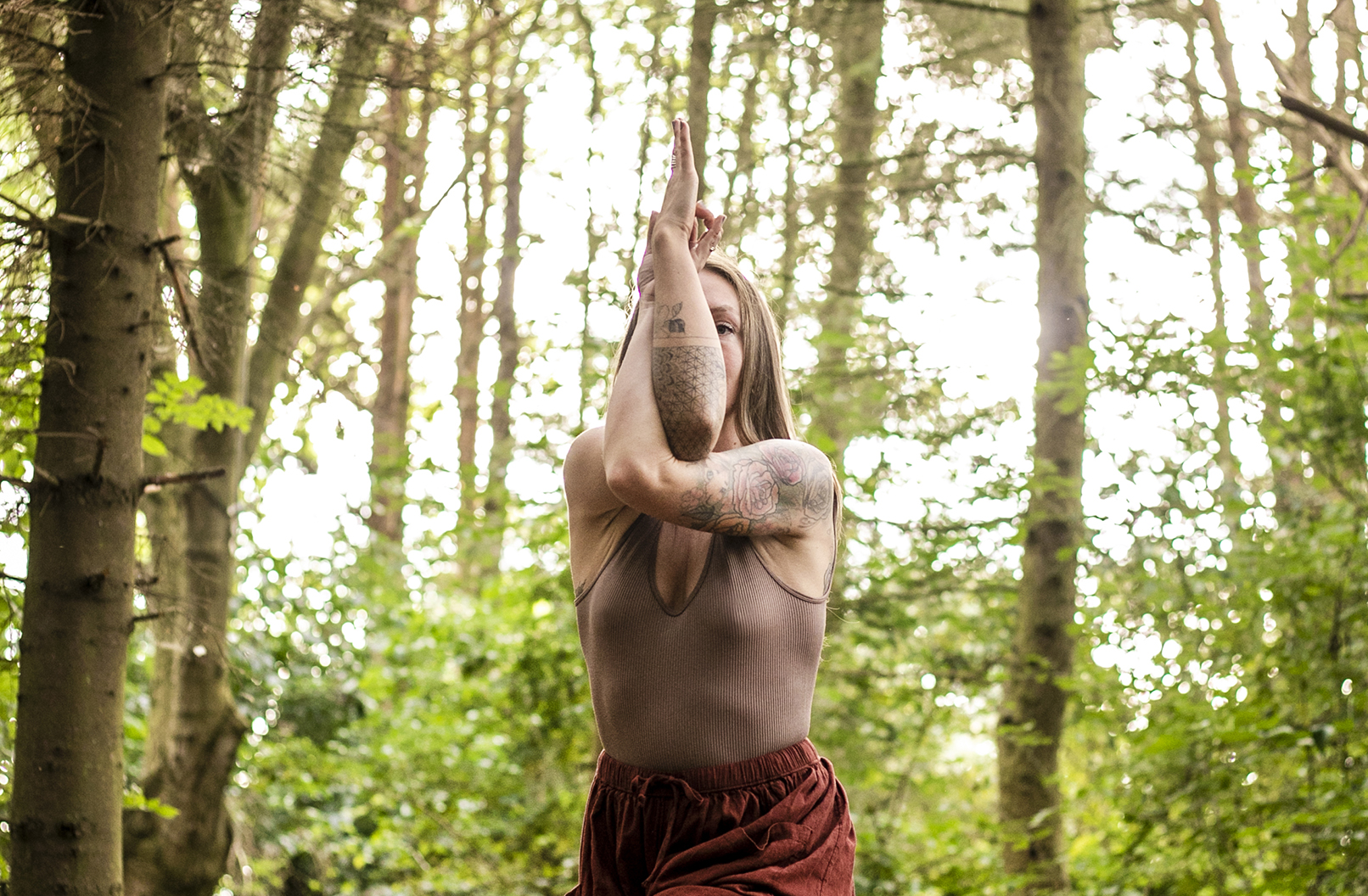 White female crossing tattooed arms over the face in a yoga position. Wearing neutral brown clothes in a woodland area