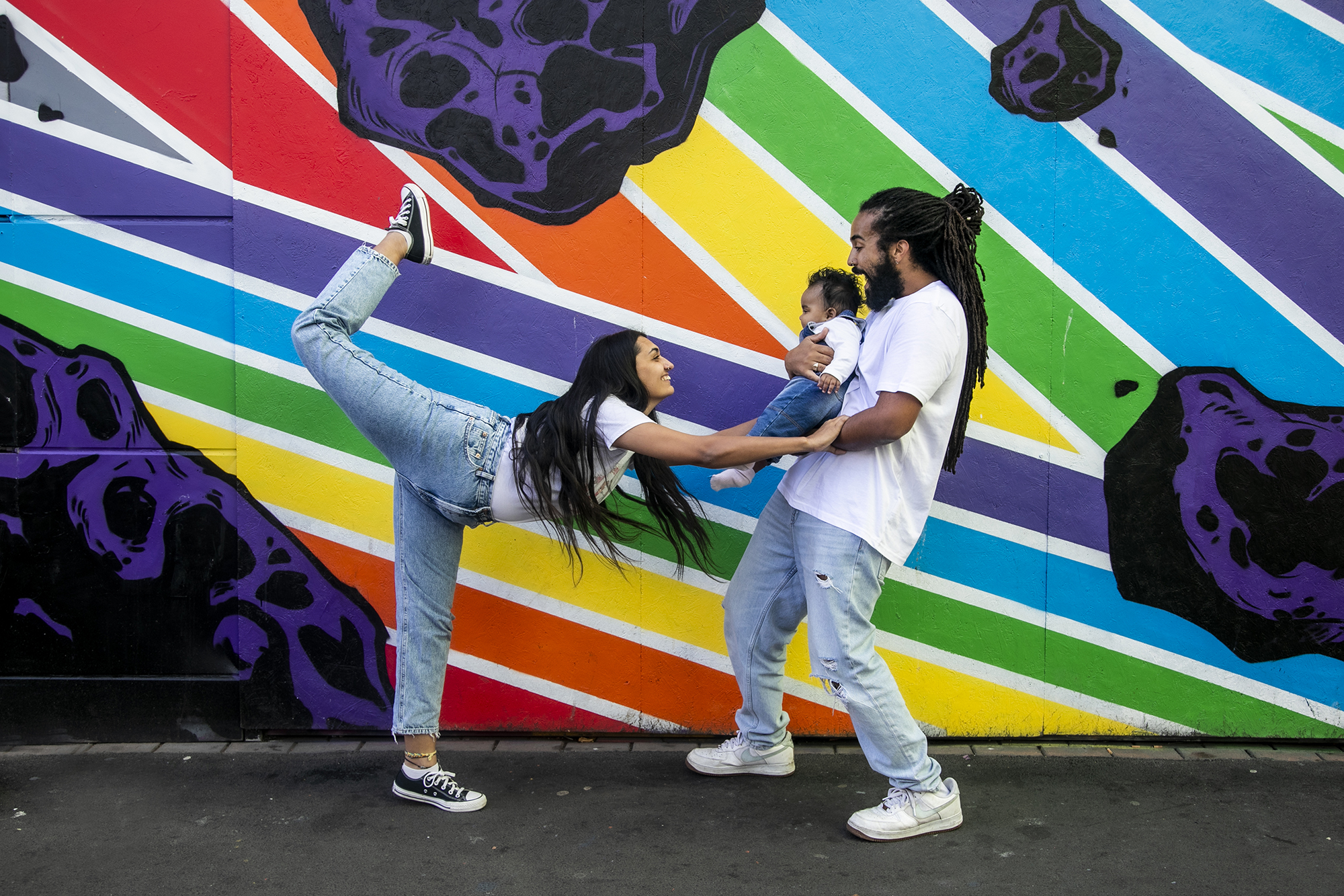 Family of 3. Global majority male with long dreadlocked hair holding baby, global majority female with long black hair, one leg up then reaching our leaning towards the baby. wearing blue jeans, trainers and white tshirts infront of rainbow graffiti wall. 