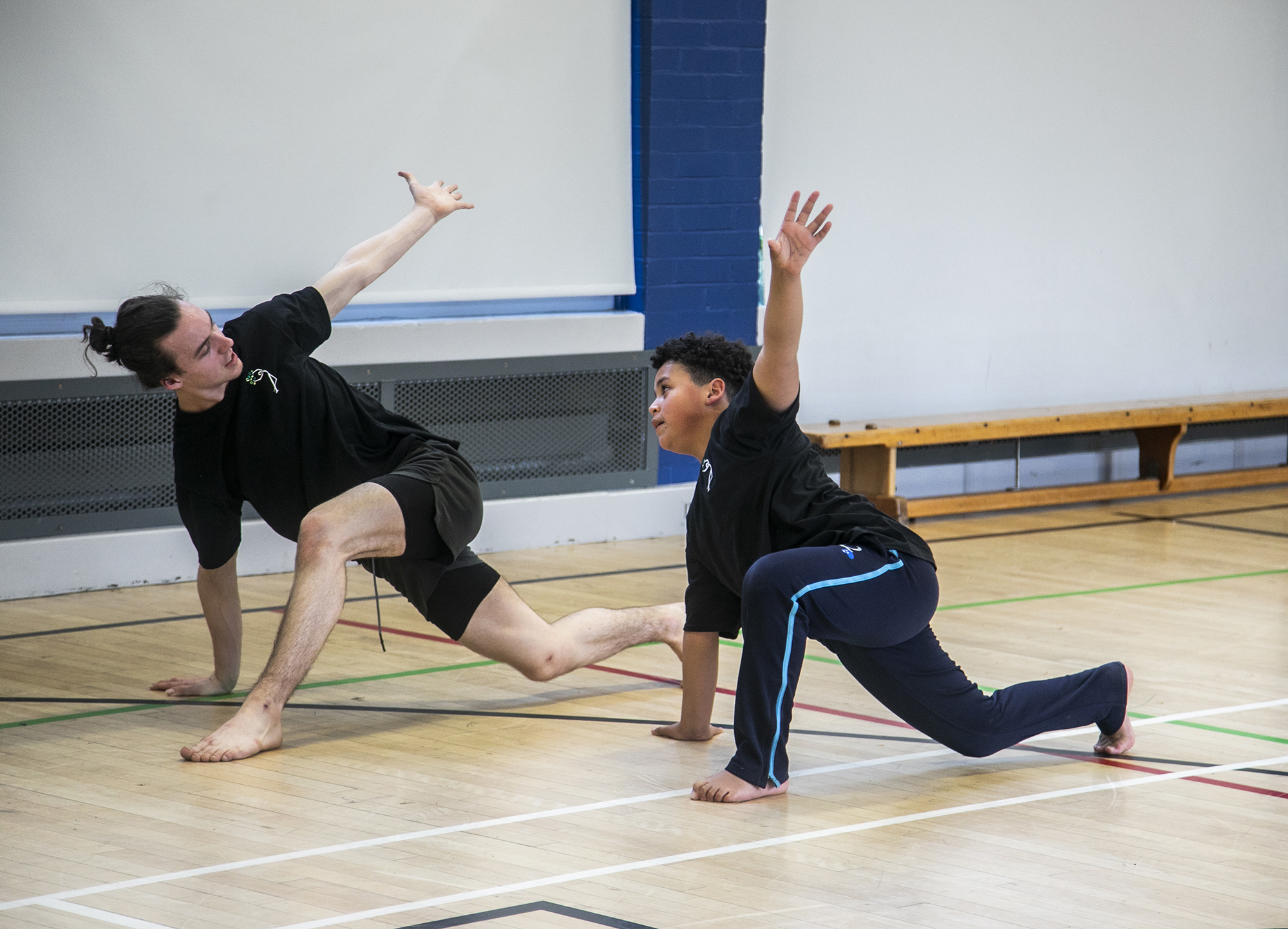 teenage male dance teacher lunging with one arm in the air teaching young boy dancer lunging with the mirrored arm in the air. In sports hall both wearing black PE kit