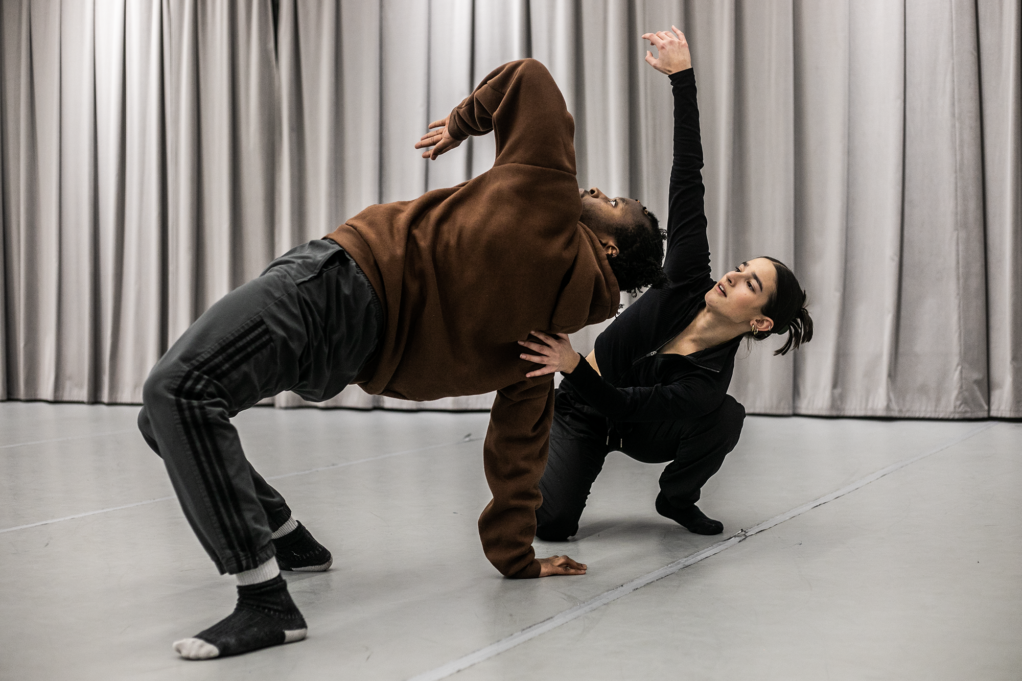 two dancers one male global majority dancer wearing brown hoodie and black joggers healing back in a bridge with one elbow pointed up. one white female wearing full black supporting other dancer with one hand and reaching up with the other