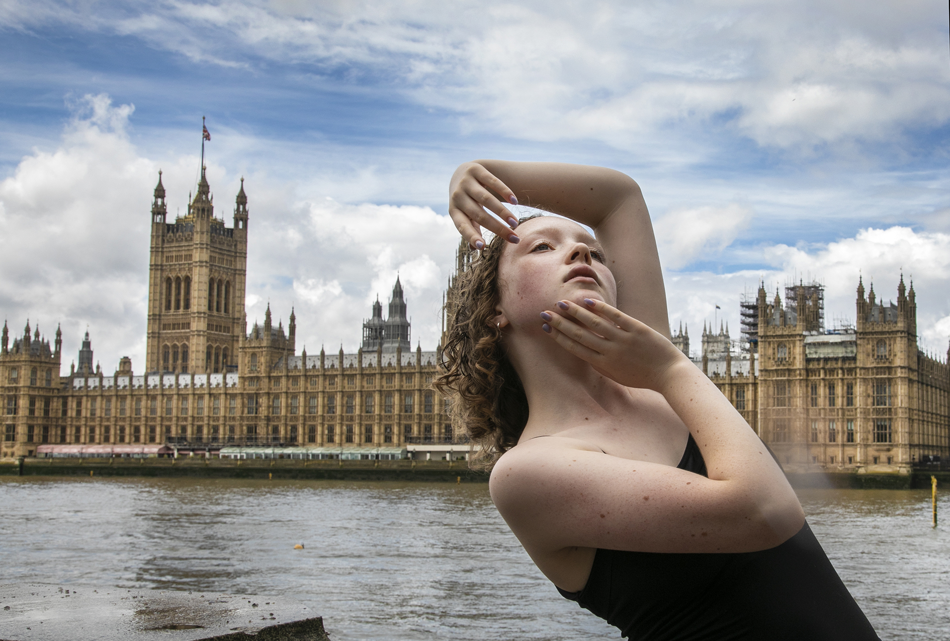 young dancer with hands around face staring up to the sky infront of the houses of parliament. White female with short curly hair and black strap top.